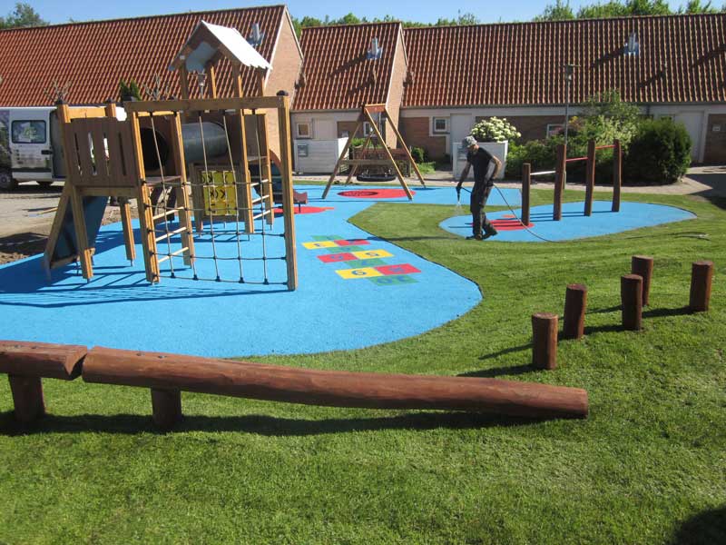 Legeområde med klatretårn, blåt gummiunderlag, træbryggeelementer og træstammer på grønt græs