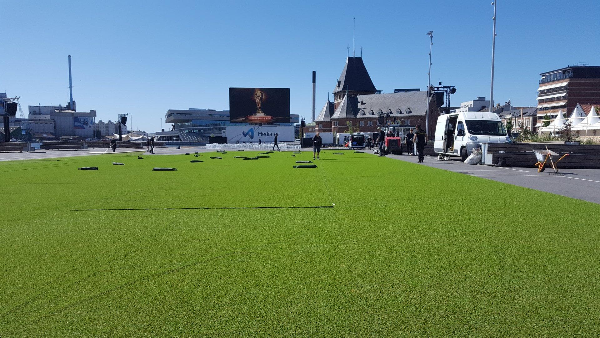 Kunstgræsanlæg under installation med rullet græs og svarte ankre på stadsplads